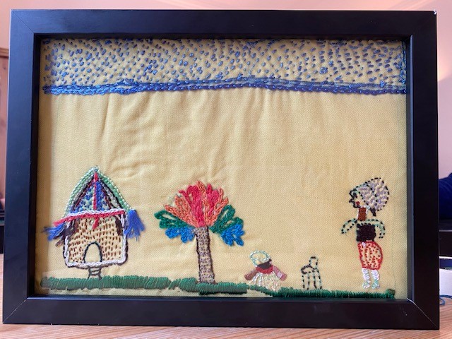 A framd textiles piece, depicting a resident's depiction of home. There is a house on the far left, a rainbow tree in the middle, and a child and a woman embroidered on the far right.