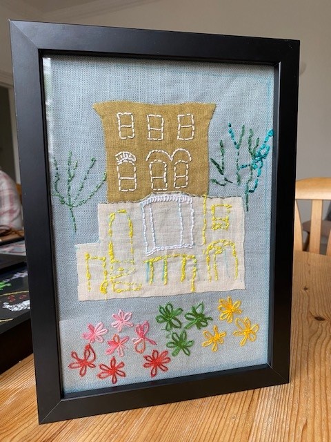 A framed embroidery piece by a resident of Emilie House, showing a depiction of Emilie House with colourful flowers in front of it.